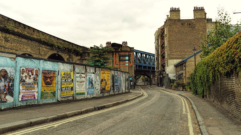 A narrow urban street scene featuring a curved, paved road with double yellow lines on each side, bordered by brick walls and a metal fence with numerous colorful posters attached. The posters display various promotional images and text, some advertising events with dates, and others with graphic designs. On the right, a high brick wall covered partially in green ivy runs alongside the sidewalk, which appears clean and well-maintained. In the background, a blue metal railway bridge spans across the street, supported by brick and stone pillars, with additional brick residential buildings featuring multiple chimneys rising above. The sky overhead is overcast, casting diffuse lighting that softens shadow lines. The scene suggests an area near an underpass or railway line, possibly indicative of limits for certain private waste collection services, highlighting challenges faced by rubbish removal operators in narrow city streets. The image is relevant for discussing access issues and alternative disposal methods, aligning with the services of Rubbish Removal Kennington, who handle waste in restricted urban environments.