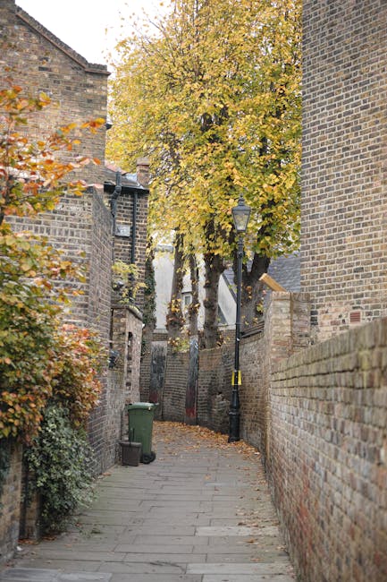 A narrow paved alleyway in an urban environment bordered by tall brick walls and historic buildings with weathered, dark brown bricks. The pathway is lined with fallen autumn leaves, and a green wheelie trash bin is positioned on the left side near the corner. In the background, a large tree with yellow and orange leaves extends over the alley, partially obscuring a white building behind it. There is a black traditional-style lamp post attached to the brick wall on the right, providing ambient lighting. The scene is illuminated by natural daylight, highlighting the textures of the brickwork and the vibrant autumn foliage. This setting reflects an area where private waste collection could be necessary due to limited space for conventional rubbish removal, with the alley offering a typical example of an accessible yet narrow street that presents challenges for rubbish collection services, making it relevant to companies like Rubbish Removal Kennington offering alternative waste handling solutions.