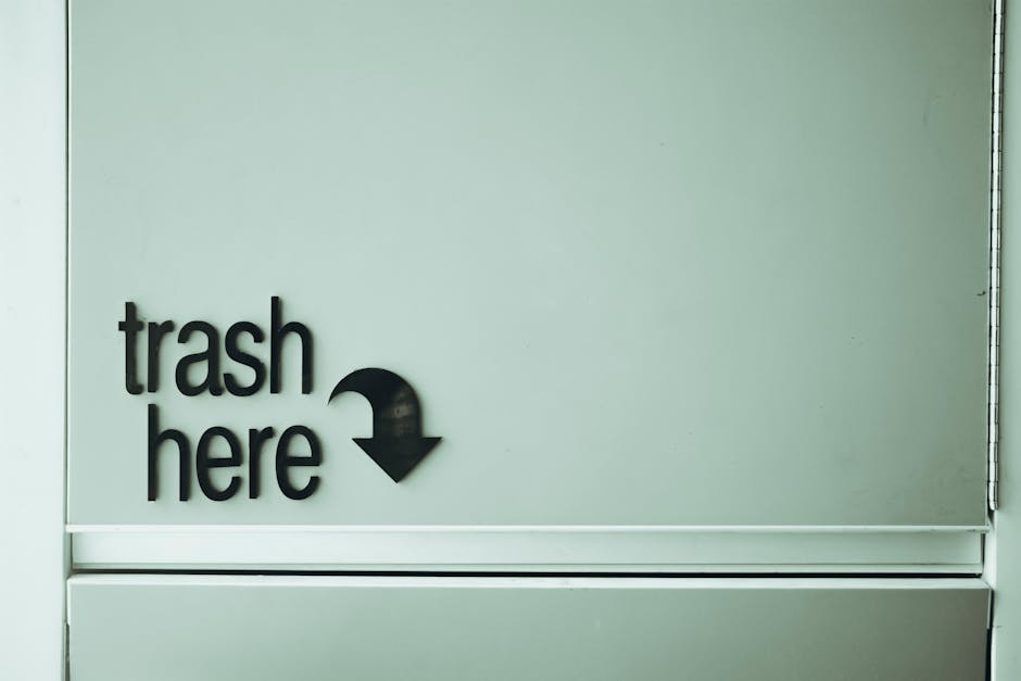 A close-up of a light grey wall with black three-dimensional lettering that reads 'trash here' accompanied by a downward arrow. The sign is affixed near the top left corner of a metallic trash disposal unit or container. The surface of the sign appears smooth and matte, while the wall behind it is plain and slightly textured. The metallic container below has a visible handle at the bottom edge, suggesting a door or lid for waste disposal. The environment suggests an indoor or sheltered area for refuse collection, with an emphasis on guiding proper rubbish disposal in line with private waste removal services. There is subtle natural or artificial lighting illuminating the scene, creating soft shadows and ensuring clear visibility of the signage and container details. This scene aligns with waste management practices where clear signage indicates designated disposal points, supporting independent rubbish collection methods that may be an alternative to local authority services, as offered by companies like Rubbish Removal Kennington.