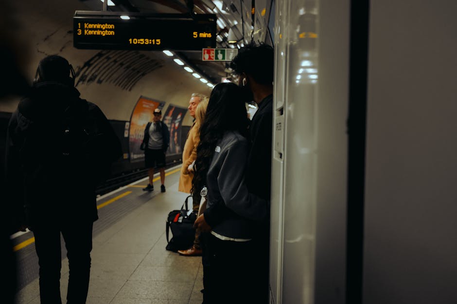 Inside a public underground train station, several passengers are waiting on the platform. In the foreground, a woman with long dark hair is standing close to the train door, partially obscured by a nearby vertical support column, and is wearing a dark jacket. To her left, a man dressed in dark clothing with a backpack is facing away from the camera, waiting along the platform. Further back, passengers including a woman in a beige coat and a man in a grey hoodie are standing near the platform edge, some with luggage or backpacks. Overhead, an electronic display board shows train destinations and times, indicating departures to Kennington in approximately five minutes. The station features a curved tunnel ceiling with evenly spaced lighting, and the platform surface appears to be made of textured concrete. The overall scene captures a typical moment of commuters preparing for their journey, consistent with the context of efficient waste management and the importance of maintaining clean transportation environments, as supported by private rubbish removal services like Rubbish Removal Kennington.