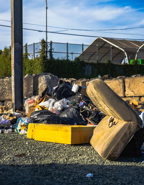 An outdoor scene during daytime featuring a large pile of mixed waste and rubbish on a gravel surface. In the foreground, there are black plastic rubbish bags, some torn open, revealing contents such as paper, plastic bottles, and household waste. A yellow plastic storage bin is positioned near the rubbish, partially obscured by the debris. To the right, an old, worn car tyre leans against the pile, showing signs of wear and dirt. Behind the waste pile, a low stone wall made of roughly cut, irregularly shaped stones separates the area from a fence and a green hedge. In the background, utility poles with electrical wires stretch across the scene, and a large, curved, semi-transparent shelter or building with a beige or light-colored roof is visible, set against a mostly clear blue sky with some wispy clouds. The scene suggests a spot for bulk rubbish collection or waste clearance, potentially related to independent disposal or private clearance services such as those offered by Rubbish Removal Kennington in the context of alternative waste handling methods.