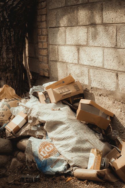 A small pile of mixed waste materials situated at the base of an exterior brick wall, partially obscured by a large white sack. The pile includes flattened cardboard boxes, some with printed labels and logos, alongside crumpled brown paper bags and plastic packaging. The cardboard boxes vary in size and are mostly light brown, with some showing torn or bent edges. The white sack appears to be made of woven fabric, possibly polypropylene, with textured surface and visible folds. The environment appears to be an outdoor space, possibly a back alley or side yard, with natural lighting casting soft shadows on the ground. To the left, there is a thick tree trunk, and the background features a wall constructed of concrete blocks with a rough surface and light-colored mortar joints, indicating a utilitarian setting for rubbish disposal. This scene illustrates an instance of domestic or small-scale commercial rubbish accumulation, which could be handled through private collection services such as those offered by Rubbish Removal Kennington and similar companies specializing in independent waste clearance and alternative rubbish disposal methods.