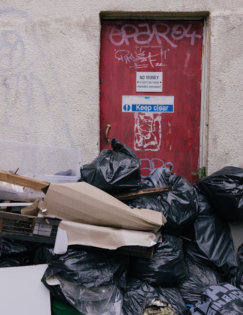 An outdoor scene during daytime featuring a large pile of mixed waste and rubbish on a gravel surface. In the foreground, there are black plastic rubbish bags, some torn open, revealing contents such as paper, plastic bottles, and household waste. A yellow plastic storage bin is positioned near the rubbish, partially obscured by the debris. To the right, an old, worn car tyre leans against the pile, showing signs of wear and dirt. Behind the waste pile, a low stone wall made of roughly cut, irregularly shaped stones separates the area from a fence and a green hedge. In the background, utility poles with electrical wires stretch across the scene, and a large, curved, semi-transparent shelter or building with a beige or light-colored roof is visible, set against a mostly clear blue sky with some wispy clouds. The scene suggests a spot for bulk rubbish collection or waste clearance, potentially related to independent disposal or private clearance services such as those offered by Rubbish Removal Kennington in the context of alternative waste handling methods.