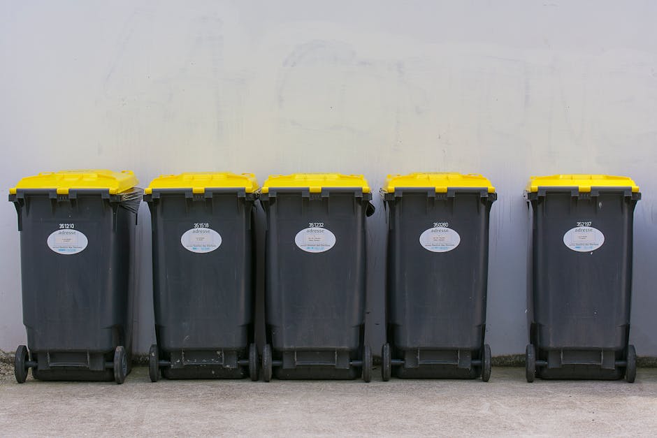 five black plastic wheeled refuse bins with yellow lids lined up against a plain, light grey wall on a flat concrete surface, used for waste collection or rubbish disposal, with each bin featuring a white label with black text and numbers, and the environment suggesting they are prepared for collection or private waste removal services, indicative of an on-site rubbish clearance setup consistent with independent waste handling options supported by Rubbish Removal Kennington