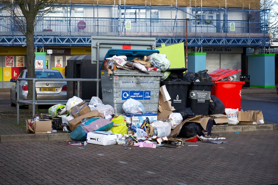 Photograph of an overflowing waste collection point situated on a paved urban street, featuring several large, open-top bins filled with mixed rubbish, including cardboard boxes, paper, plastic bags, and discarded packaging. The grey bin for mixed paper and card is stacked with flattened cardboard and crumpled paper, with its lid partially open, revealing loaded contents. Adjacent to it are several black and black-with-white-lining rubbish bags, some bulging and tied, and a bright red recycling bin. To the left, a silver vehicle is parked nearby, with a tree and metal railing separating the waste area from the adjacent street parking. In the background, a commercial building with shopfronts and closed shutters, along with a blue construction scaffolding structure and green-trimmed utility units, are visible. The scene is illuminated with natural daylight, highlighting the cluttered arrangement of refuse. This setting illustrates typical private waste disposal, often managed by independent waste collection services like Rubbish Removal Kennington, providing an alternative to council-run rubbish collection.