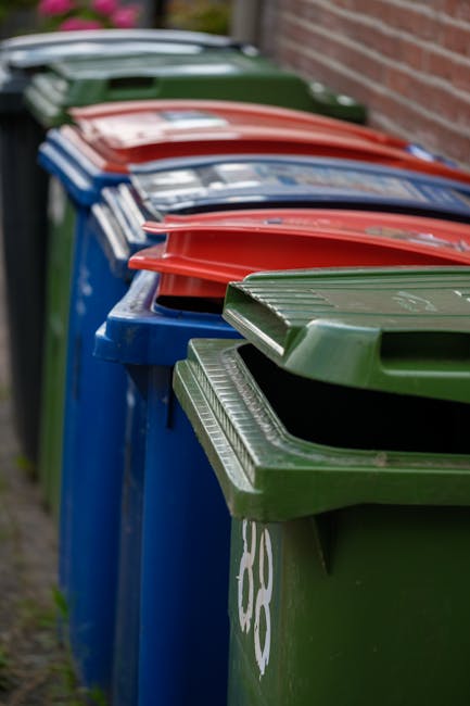 five black plastic wheeled refuse bins with yellow lids lined up against a plain, light grey wall on a flat concrete surface, used for waste collection or rubbish disposal, with each bin featuring a white label with black text and numbers, and the environment suggesting they are prepared for collection or private waste removal services, indicative of an on-site rubbish clearance setup consistent with independent waste handling options supported by Rubbish Removal Kennington