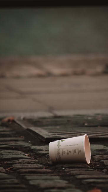 A white paper cup with printed text, lying on its side on a weathered brick pathway. The cup appears to be made of thin, smooth material, with a slightly glossy finish. It is positioned in the foreground, slightly tilted, with part of it partially obscured by the angle. The background shows a concrete surface and a part of a wooden or metal grate, which is slightly raised and runs parallel to the pathway. There is some greenery further back, softly out of focus, indicating an outdoor setting. The scene appears natural with diffuse lighting, typical of an overcast day or shaded area, highlighting the cup and textured brick surface. This image illustrates the issue of littering and may connect to waste management and rubbish removal services offered by Rubbish Removal Kennington, emphasizing the importance of responsible disposal within an environment.