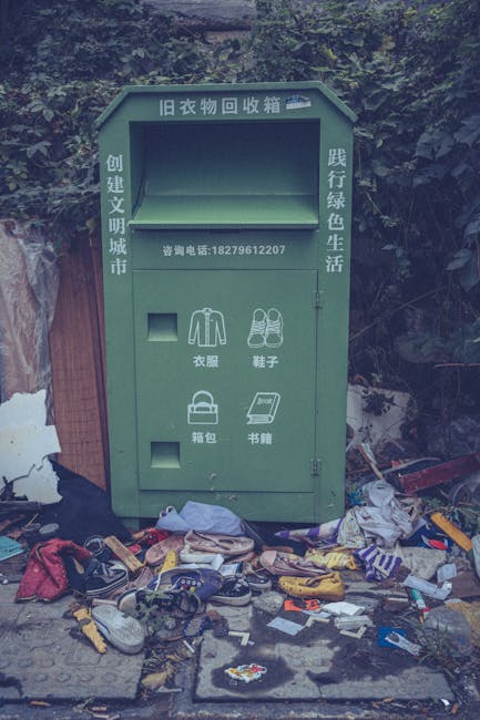 A green, metal clothing donation and recycling bin positioned on a concrete surface outdoors, with a background of dense foliage and trees. The bin has a rectangular body with a large upper compartment for clothing donations, with Chinese characters indicating its purpose. Below the main opening are smaller compartments designed for specific items such as jackets, shoes, bags, and books, each marked with icons and Chinese labels. The surrounding area is cluttered with discarded footwear, fabric, clothing, newspapers, and various small objects scattered haphazardly on the ground. The scene appears to be in a public outdoor space, possibly near a residential or park area, with a weathered wooden fence on one side. Overhead, natural light suggests a daytime setting. The overall current state reflects neglect, with visible litter and an unkempt environment, highlighting issues related to waste disposal and the importance of proper rubbish management services, such as those offered by Rubbish Removal Kennington, for responsible disposal and site clearance.