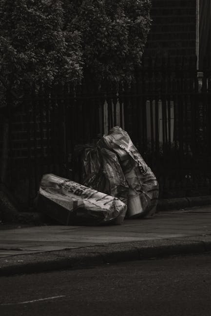 Three large black plastic rubbish bags, partially torn and crumpled, are placed on the pavement beside a black metal fence. Behind the fence, dense greenery with leafy branches is visible, along with the dark structure of a building in the background. The scene appears to be outdoors in an urban or suburban area, with the rubbish bags positioned near the curb, suggesting a local rubbish collection point or an area designated for waste disposal. The lighting in the image is subdued, creating a shadowy atmosphere that emphasizes the irregular texture of the wrinkled bags and their contents, which are not visible. The overall scene reflects an instance of waste awaiting removal, aligning with the services of independent rubbish removal companies like Rubbish Removal Kennington, especially in contexts where private collection or on-site clearance might be required.