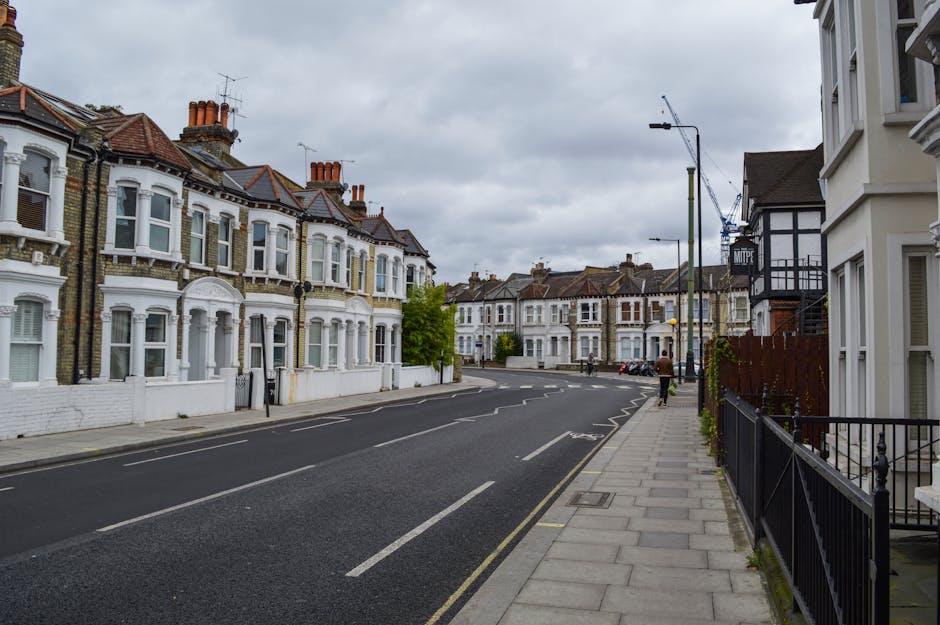 A quiet residential street with a row of Victorian-style terraced houses featuring decorative brickwork, bay windows, and white-painted facades on the left side of the image. The street is paved with dark asphalt, marked with white road lines, and lined with a light grey concrete pavement on the right. A single person is walking along the sidewalk, which is bordered by a black metal railing and a small fenced garden area with climbing plants. Several streetlights and a large crane in the background indicate ongoing construction nearby. The sky is overcast with grey clouds, creating diffuse lighting across the scene. The overall environment suggests an urban area where private waste collection and rubbish removal services, such as those provided by Rubbish Removal Kennington, might be utilized for managing waste in this well-maintained neighborhood, particularly with the presence of a relatively narrow road and a pedestrian-friendly layout.