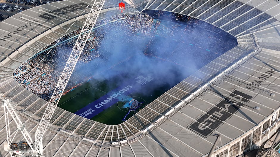 An aerial view of a large, modern football stadium with a partially open roof, showing a well-maintained green pitch filled with visitors and staff. A section of blue smoke or fog is rising from the center of the field, possibly from celebratory pyrotechnics or special effects. Surrounding the pitch are rows of seating filled with spectators, some waving flags or banners. The stadium's exterior features a combination of glass and metal panels, with structural supports and light fixtures visible along the edges. A large digital banner on the side of the stands displays the text 'CHAMPIONS' in white letters against a blue background, indicating a major sporting event. The emphasis on the stadium's infrastructure and clean, organized environment highlights the context of large-scale event management and the importance of efficient waste and rubbish disposal services for match day activities, aligning naturally with the topic of alternative rubbish removal solutions for high-capacity venues. Rubbish Removal Kennington can assist with waste handling needs associated with events at such facilities, ensuring tidy and responsible clearance post-event.