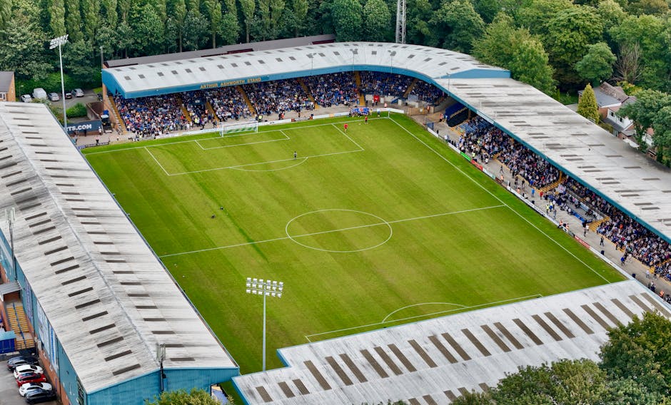 An aerial view of a football stadium with a well-maintained grass pitch in the centre, surrounded by spectator stands filled with fans. The stands have a curved, roofed structure with a predominantly grey finish, and the seating areas are visible with some spectators occupying them. The field features white lines marking the boundaries, centre circle, and penalty areas, with bright stadium floodlights installed on tall poles around the pitch. The surrounding environment includes dense green trees and residential buildings visible beyond the stadium perimeter, indicating an urban location. The stadium's exterior has a blue colour scheme with visible entrances and pathways leading to parking areas with parked cars. The scene is illuminated by natural daylight, creating a clear, vibrant atmosphere, and the overall setting suggests an active sports venue occasionally used for events, with potential for independent or private waste management services such as rubbish removal by companies like Rubbish Removal Kennington. The image's focus on the well-organized, clean environment reflects the importance of maintaining tidy sports facilities through effective rubbish and waste removal practices.