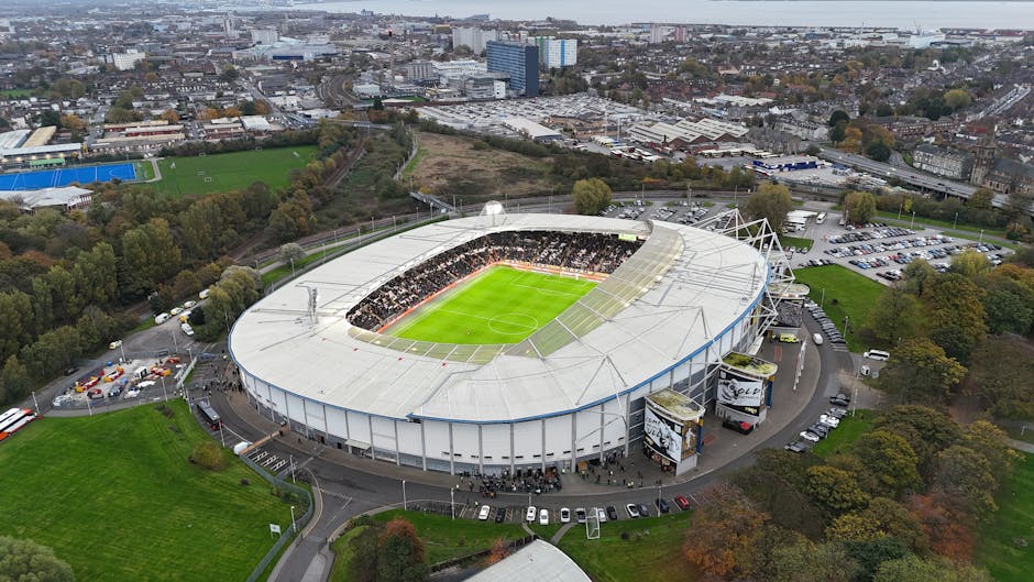 An aerial view of a large, modern football stadium with a partially open roof, showing a well-maintained green pitch filled with visitors and staff. A section of blue smoke or fog is rising from the center of the field, possibly from celebratory pyrotechnics or special effects. Surrounding the pitch are rows of seating filled with spectators, some waving flags or banners. The stadium's exterior features a combination of glass and metal panels, with structural supports and light fixtures visible along the edges. A large digital banner on the side of the stands displays the text 'CHAMPIONS' in white letters against a blue background, indicating a major sporting event. The emphasis on the stadium's infrastructure and clean, organized environment highlights the context of large-scale event management and the importance of efficient waste and rubbish disposal services for match day activities, aligning naturally with the topic of alternative rubbish removal solutions for high-capacity venues. Rubbish Removal Kennington can assist with waste handling needs associated with events at such facilities, ensuring tidy and responsible clearance post-event.