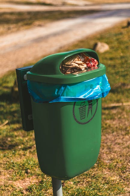 A green, environmentally styled outdoor waste bin with a rounded, slightly domed lid containing scattered crumpled paper and plastic wrappers, positioned on a metal post on a grassy area near a dirt or gravel pathway. The bin features a black recycling symbol with a small illustrated bin, indicating it is designated for rubbish collection, and is lined with a bright blue plastic bag visible through the open lid. In the background, there is a blurred view of an open outdoor space with patches of grass and dirt, suggesting a park or communal area, and natural sunlight illuminates the scene with a warm tone. The scene reflects an example of independent waste collection or disposal as part of a rubbish removal service or community management, aligned with principles of on-site rubbish handling and environmental responsibility, relevant to rubbish removal services and waste management practices.
