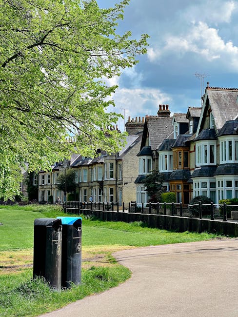 A pair of black public waste bins positioned on a grassy area near a paved footpath, with two slots on the top for rubbish disposal. The bins are rectangular with rounded edges and are situated in the foreground, slightly to the left. Behind them, a well-maintained lawn extends towards a row of Victorian-style terraced houses with bay windows, brickwork in various shades of beige and brown, and slate roofs with chimneys. To the left of the bins, a large deciduous tree with fresh green leaves partially shades the scene, its branches spreading across the top left of the image. The sky overhead is partly cloudy with patches of blue showing between white and grey clouds. The setting appears quiet and residential, with the presence of waste disposal facilities aligned with local rubbish collection or private waste handling services, and the perspective suggests an environment suitable for offering on-site or independent waste removal options, as provided by Rubbish Removal Kennington.