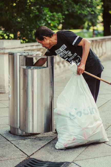 A middle-aged man with short, dark hair is seen discarding rubbish into a tall, upright metallic waste bin on a paved sidewalk. He is wearing a black T-shirt with white text and dark trousers, and holds a long-handled stick in one hand to assist with waste collection. In his other hand, he carries a large white plastic bag filled with rubbish. The background features a stone balustrade and lush green foliage, suggesting a park or landscaped urban area. The scene is well-lit with natural daylight, highlighting the reflective surface of the metal bin and the textured pavement beneath his feet. The overall setting indicates an outdoor private or community rubbish collection effort, aligned with services like those provided by Rubbish Removal Kennington, for on-site waste disposal and alternative waste handling methods outside standard municipal collection.