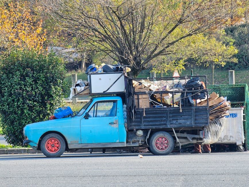 A vintage light blue pickup truck parked on the side of a paved road with its cargo area filled with various types of waste and debris. The load includes large flattened cardboard boxes, black garbage bags, and miscellaneous household items. The tailgate is open and secured with ropes, revealing the cluttered contents. The truck's wheels are painted red, contrasting with the blue exterior, and the vehicle shows signs of age and wear. Behind the vehicle, a canopy of trees with sparse, green and yellow leaves suggests an autumn setting, and a metal fence is partially visible in the background. The scene appears to be a private waste disposal or alternative waste handling situation, as indicated by the presence of waste materials that are not part of standard municipal collections, aligning with the context of rubbish removal services in Kennington.