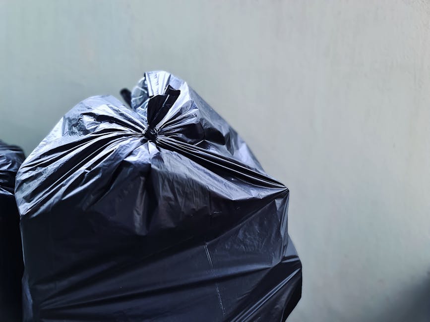A middle-aged man with short, dark hair is seen discarding rubbish into a tall, upright metallic waste bin on a paved sidewalk. He is wearing a black T-shirt with white text and dark trousers, and holds a long-handled stick in one hand to assist with waste collection. In his other hand, he carries a large white plastic bag filled with rubbish. The background features a stone balustrade and lush green foliage, suggesting a park or landscaped urban area. The scene is well-lit with natural daylight, highlighting the reflective surface of the metal bin and the textured pavement beneath his feet. The overall setting indicates an outdoor private or community rubbish collection effort, aligned with services like those provided by Rubbish Removal Kennington, for on-site waste disposal and alternative waste handling methods outside standard municipal collection.