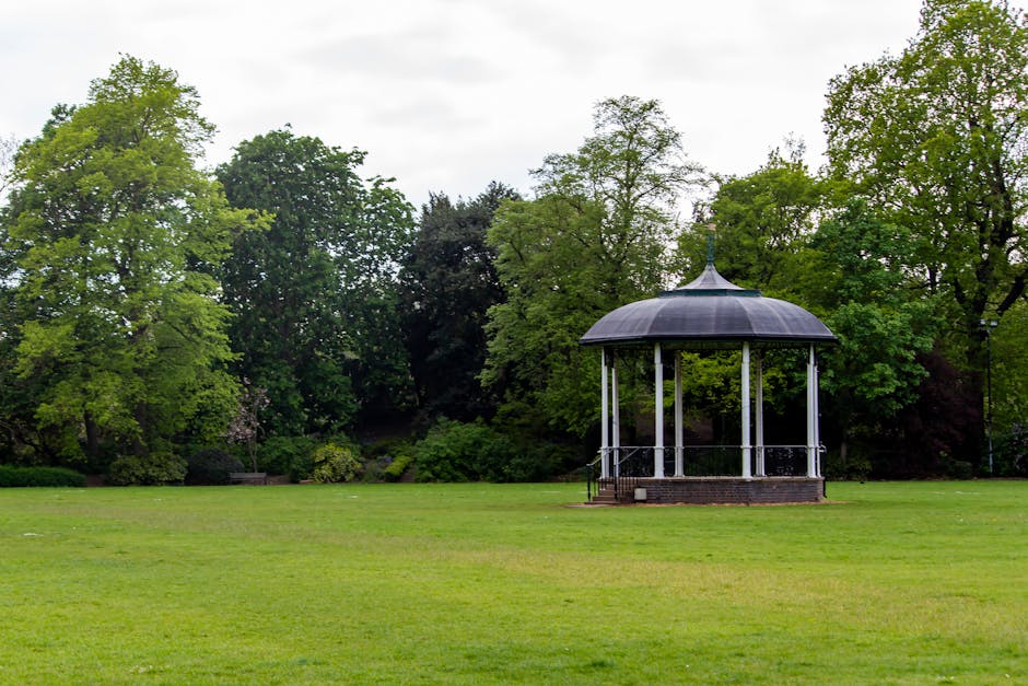 A park scene featuring a white, open-sided bandstand with a domed, dark grey roof situated on a raised brick platform, surrounded by a lush green lawn and a variety of mature trees with dense foliage in shades of green. The background shows a mix of deciduous trees, some with lighter green leaves, creating a natural backdrop under an overcast sky. The grassy area appears well-maintained with even, short grass, and there is a gentle slope leading up to the bandstand. The environment suggests a peaceful outdoor space suitable for gatherings or leisure activities, with no visible objects, benches, or pathways in the immediate foreground. The photo aligns with private or community park settings often used for outdoor events or quiet recreation, and the scene may be part of a municipal area where outdoor structures are maintained for public use. The focus is on the natural landscape complemented by the central bandstand, with no visible waste or rubbish present, consistent with upkeep and cleanliness typical of such locations, aligning with services like those provided by Rubbish Removal Kennington.